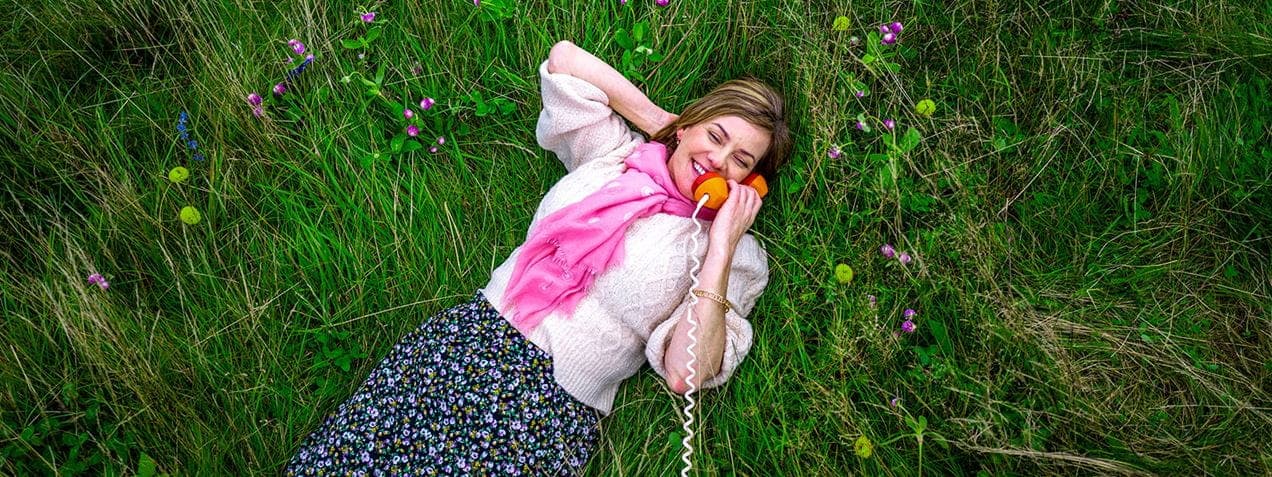 A woman lies on grass with a smile, talking on an orange telephone receiver. She wears a pink scarf, white blouse, and floral skirt. Surrounding her are small purple and yellow wildflowers, creating a serene and colorful outdoor setting.
